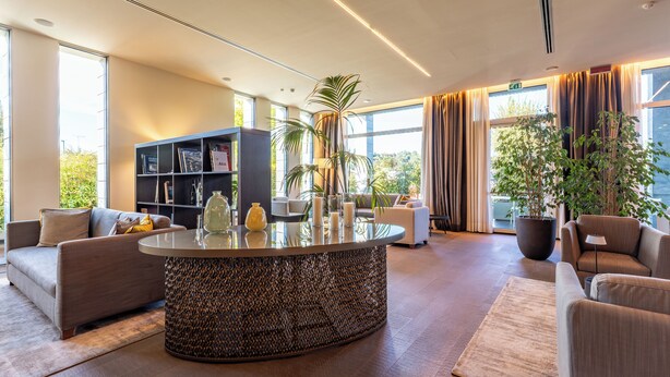 A modern hotel lobby with a central round table, sofas, a bookshelf, and large windows with curtains. - HSM Hotel San Martino (Garbagnate Monastero)