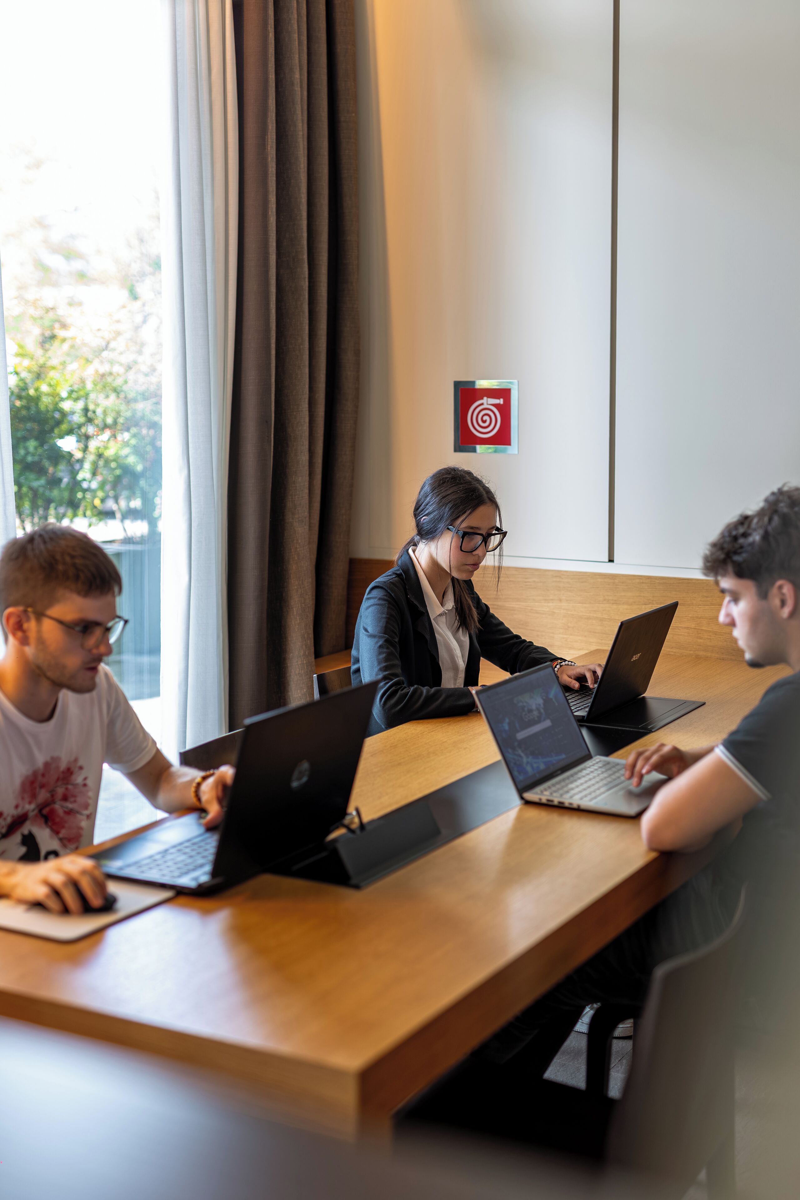 Tres personas trabajando en una mesa de madera con computadoras portátiles, en una habitación con un ventanal grande y un cartel con un blanco en la pared.