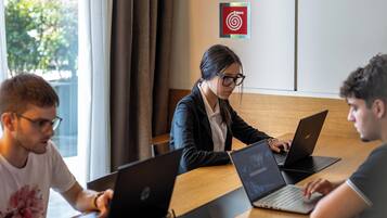 Three people working at a wooden table with laptops, in a room with a large window and a target poster on the wall.