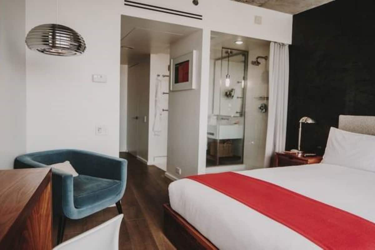Una habitación de hotel moderna con una cama grande, un escritorio, una silla azul y zona de ducha.