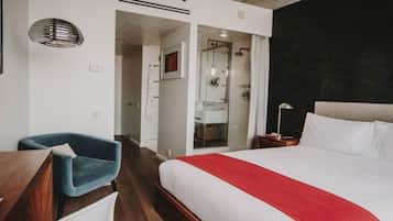 Una habitación de hotel moderna con una cama grande, un escritorio, una silla azul y zona de ducha.
