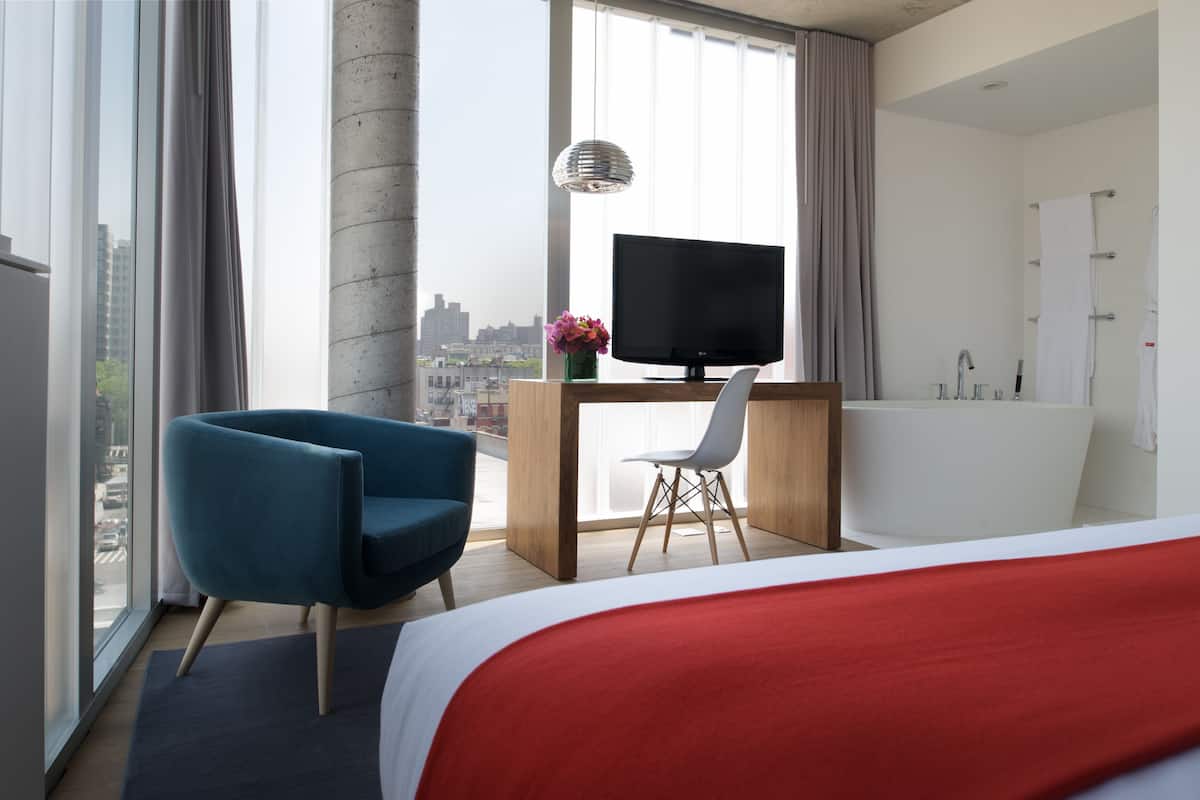 Habitación de hotel con cama, escritorio con televisor, silla, ventana grande con vista a la ciudad y bañera.