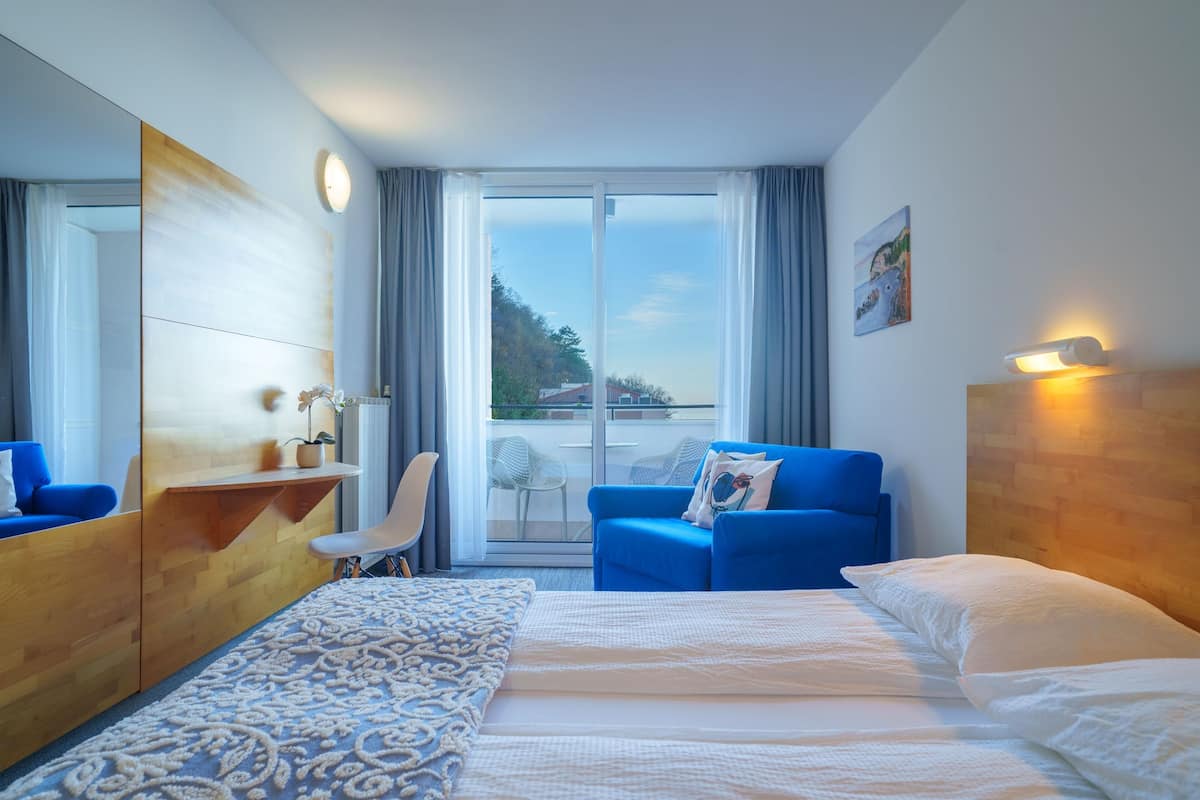 A hotel room with a large bed, a blue sofa, a wooden desk, a chair, and a balcony with a view of the outdoors.