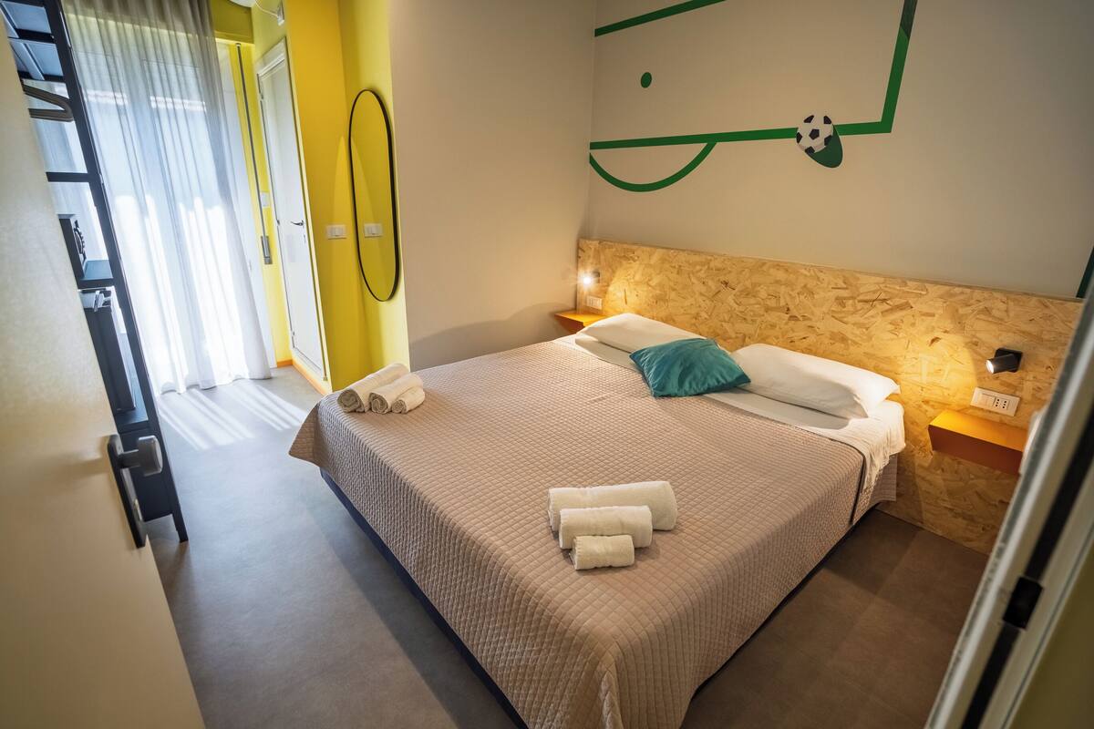 A hotel room with a bed, a headboard, a wall clock, a bedside table, and a window with curtains.