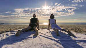 Two people stretching on a beach blanket facing the ocean with the sun setting in the background. - Fantini Club Hotel  (Cervia)