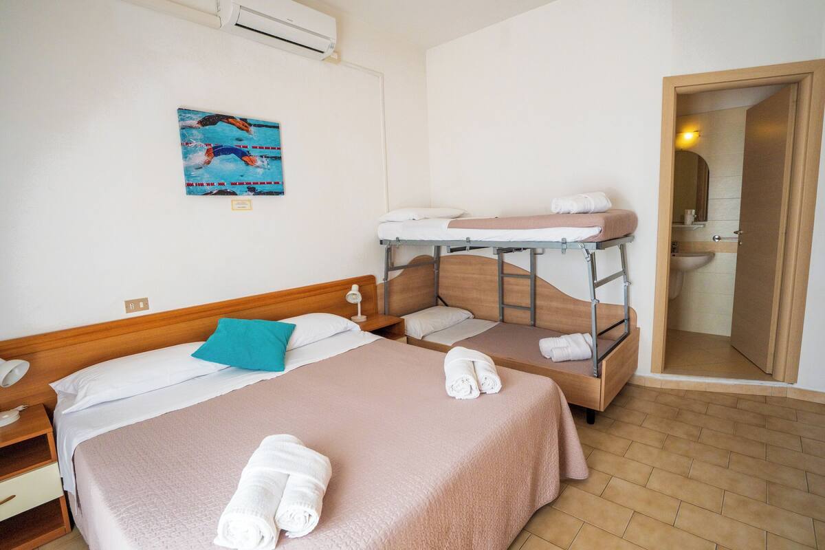 A hotel room with two beds, a bunk bed, a painting, a bedside table, a lamp, and a bathroom visible through an open door.