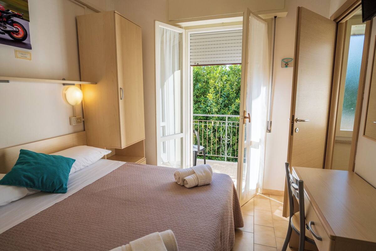 A hotel room with a bed, a desk, and a balcony with a view of greenery.