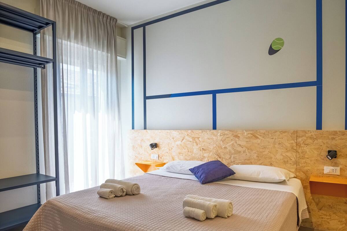 A hotel room with a bed, a nightstand, a wall-mounted lamp, and a window with curtains.