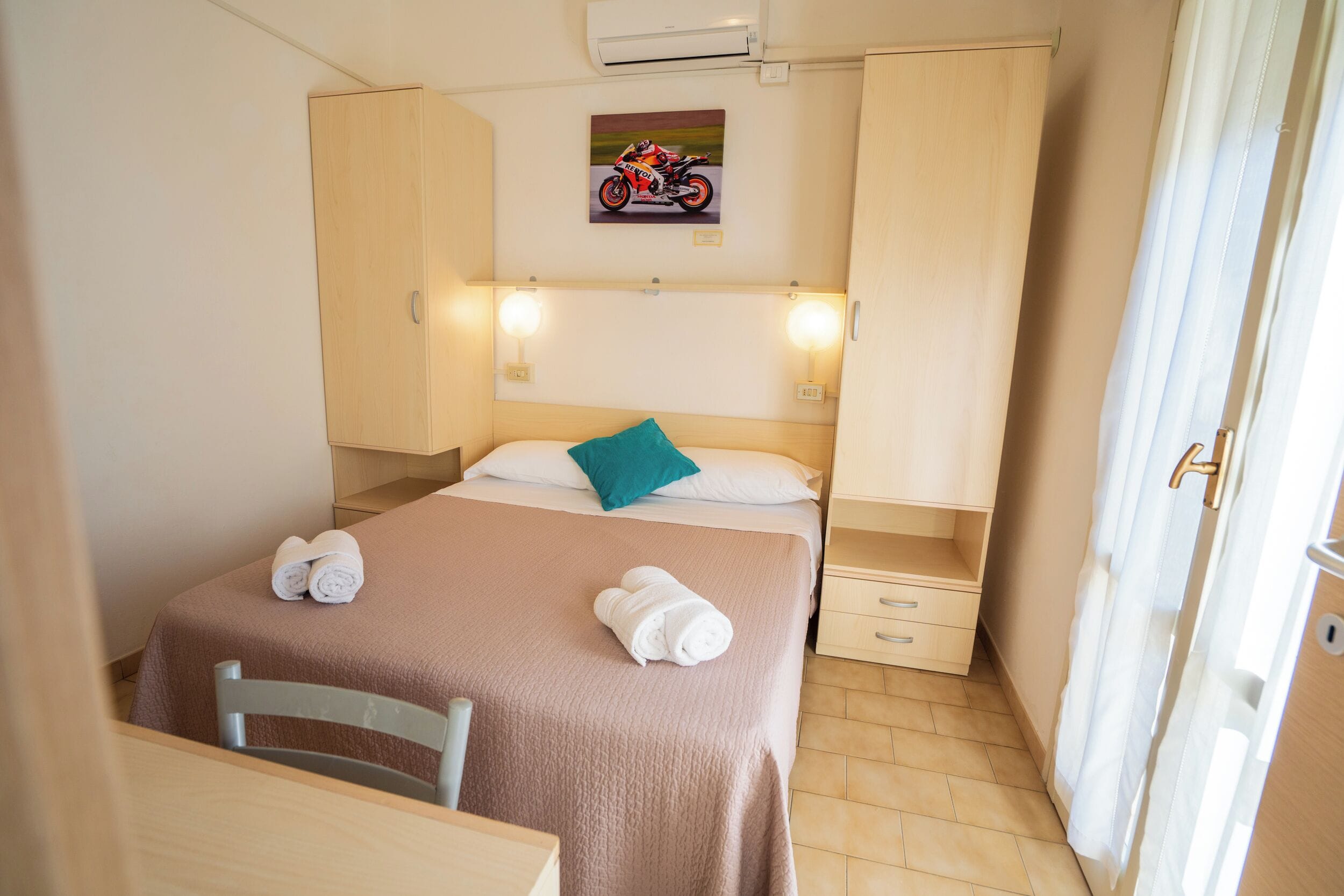 A hotel room with a bed, a chair, a small table, a nightstand, a lamp, a painting, and a window with curtains.