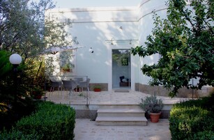 A white building with a glass door, a patio with chairs, and potted plants.