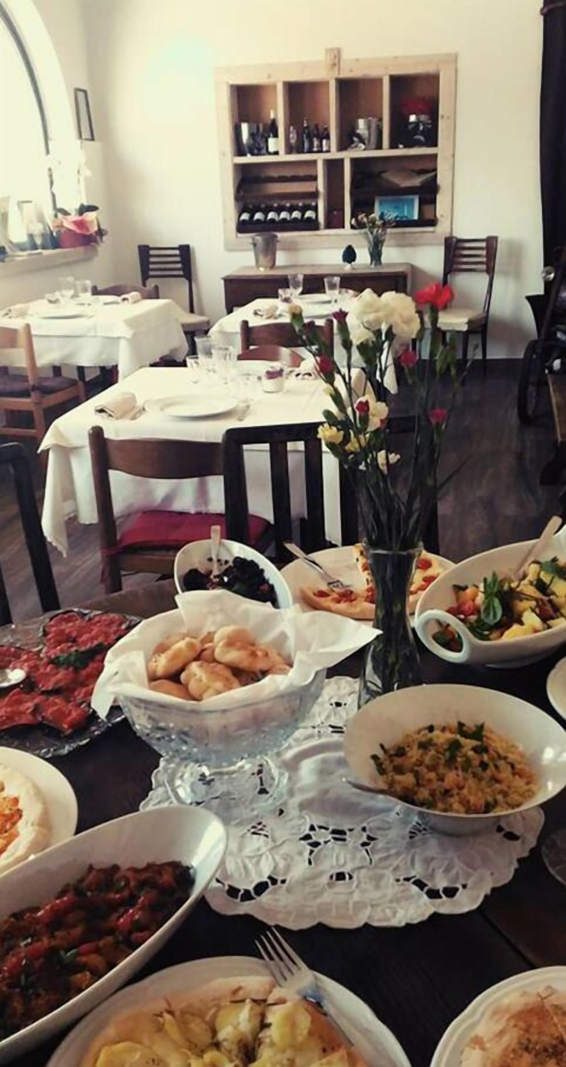 Comedor con varias mesas dispuestas para una comida, una bodega en el fondo y un jarrón con flores sobre la mesa.