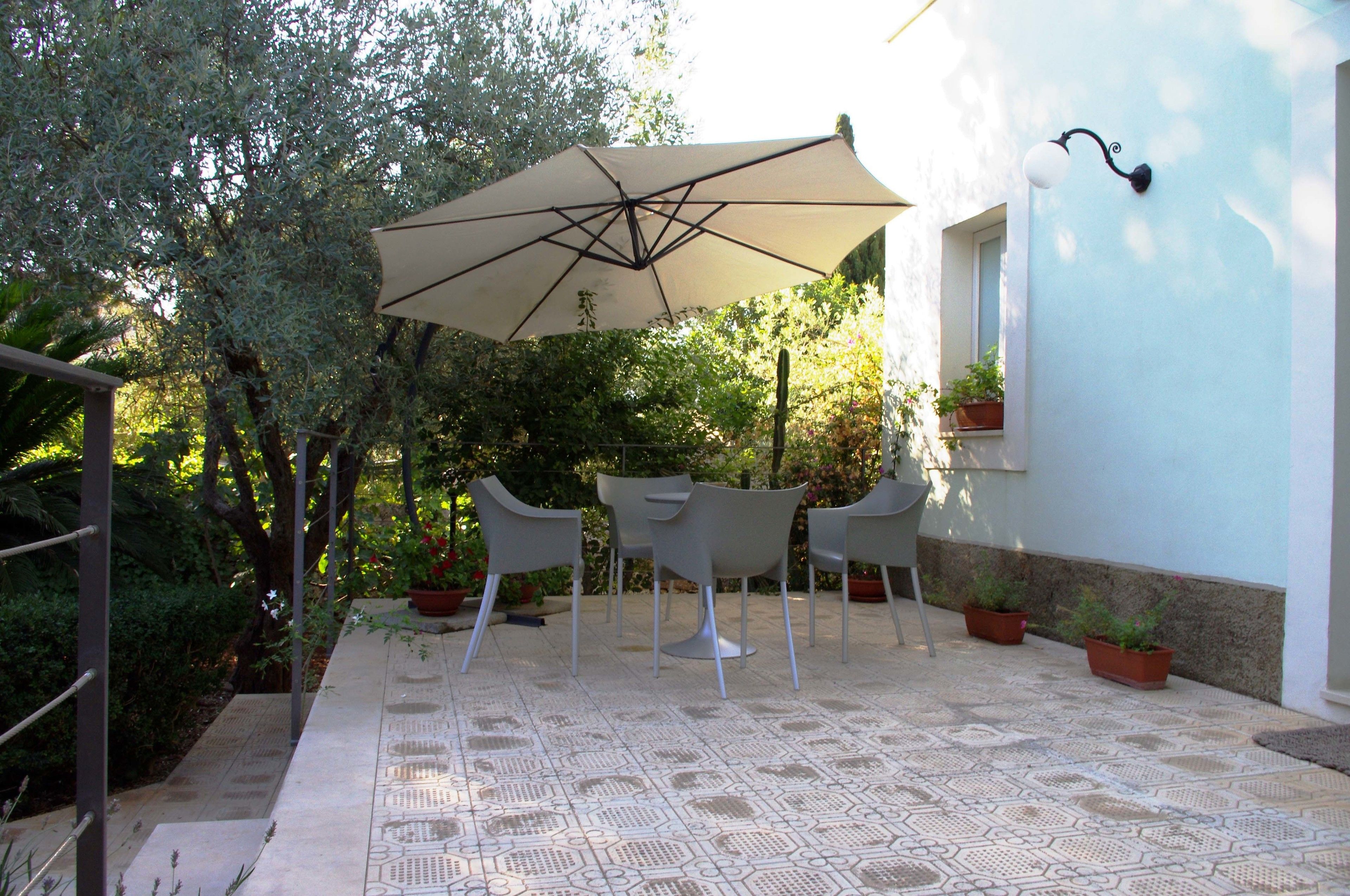 Terrassa exterior amb taula i cadires, un parasol gran i plantes en testos.