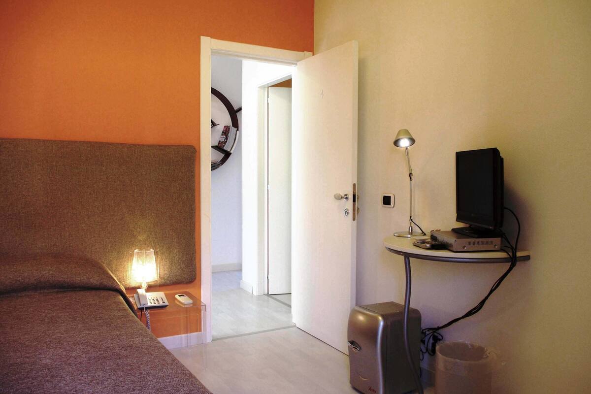 Una habitación de hotel con una cama, un escritorio con lámpara, una computadora y un cesto de basura.