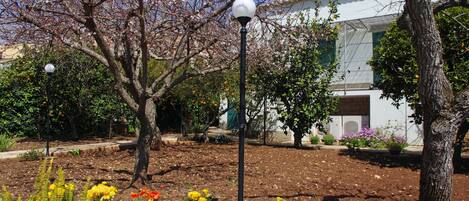 A garden with flowering plants and trees, a lamp post, and a building with a white facade and green shutters.
