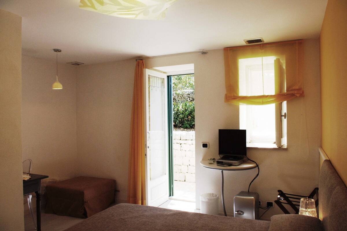 Una habitación pequeña y bien iluminada con una cama, un escritorio con computadora y una ventana con cortinas.
