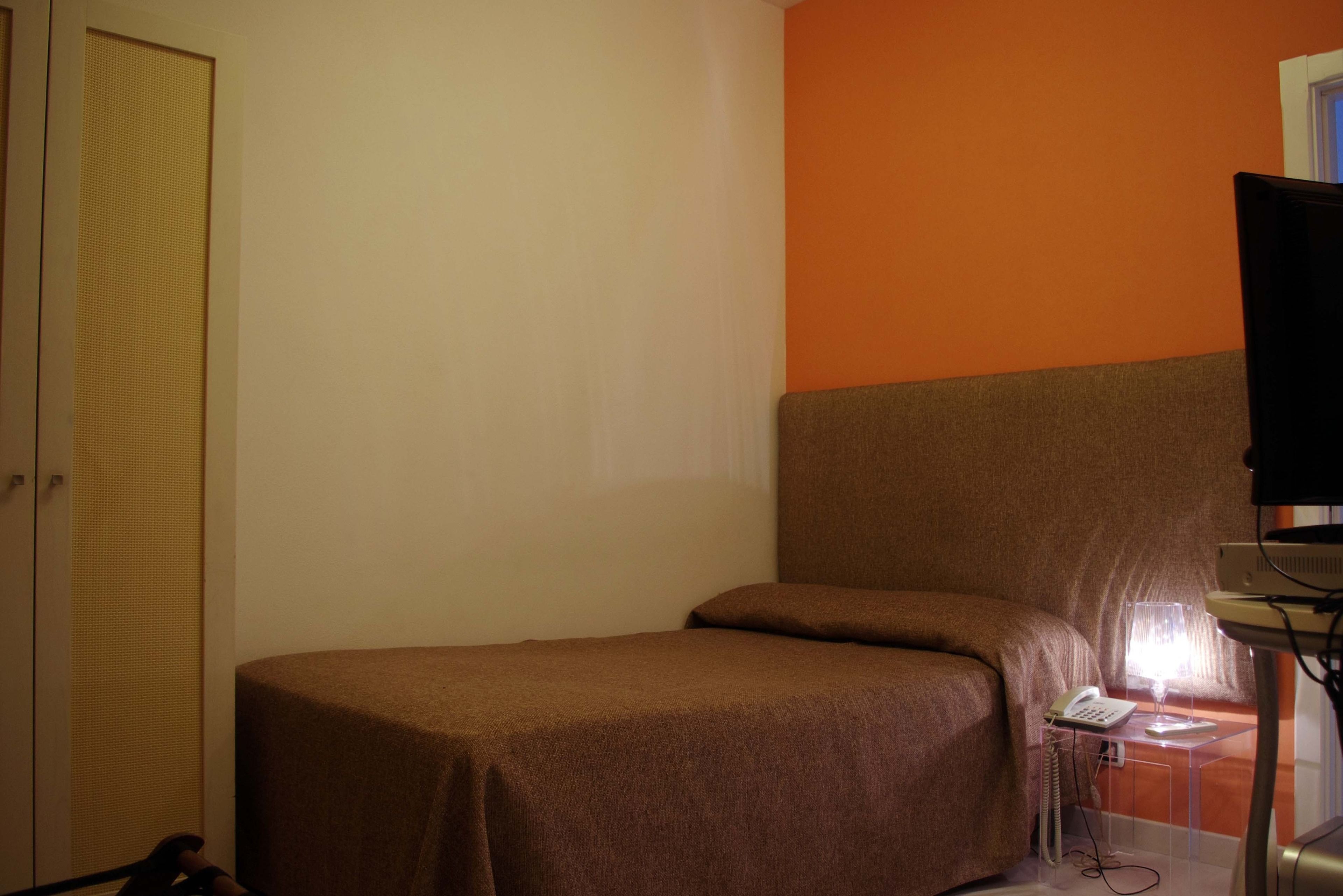 A small, single-bed room with a brown bedspread, a bedside telephone, and a lamp on a small table.