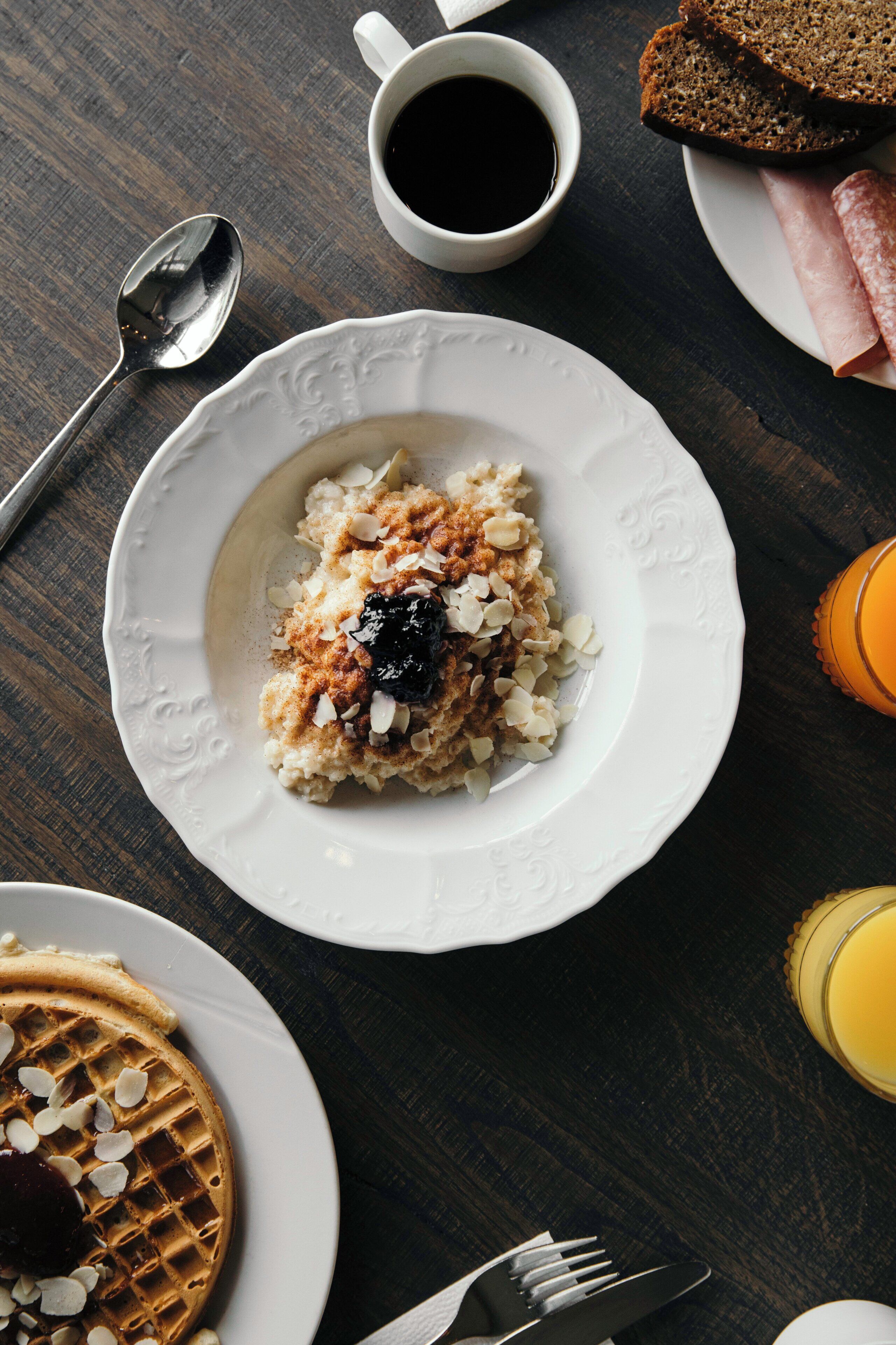Desayuno con avena, tostadas, jamón, un gofre y una taza de café sobre una mesa de madera.