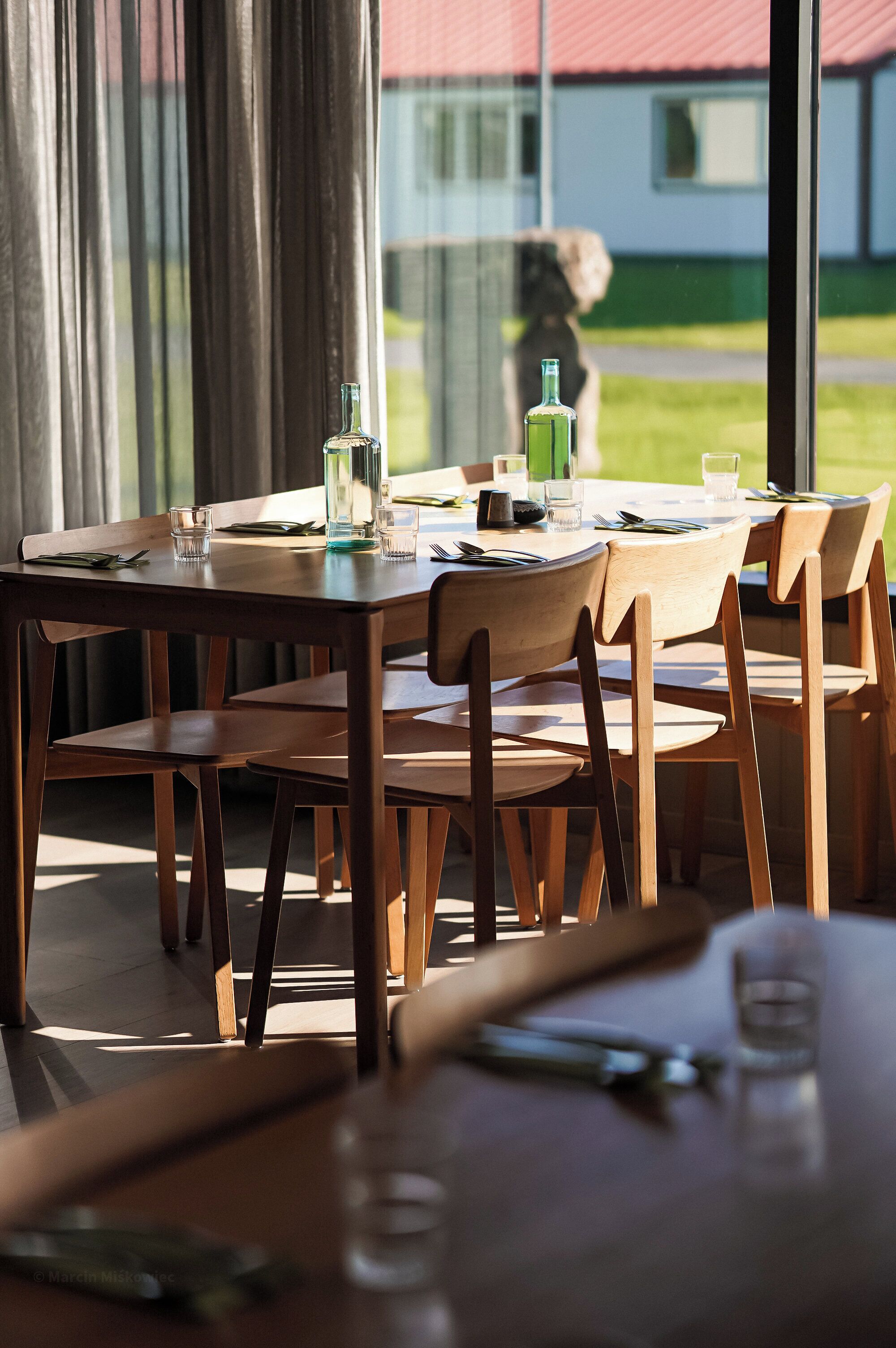 Mesa de comedor dispuesta con sillas, una botella y vasos, con vista a un edificio y follaje exterior.
