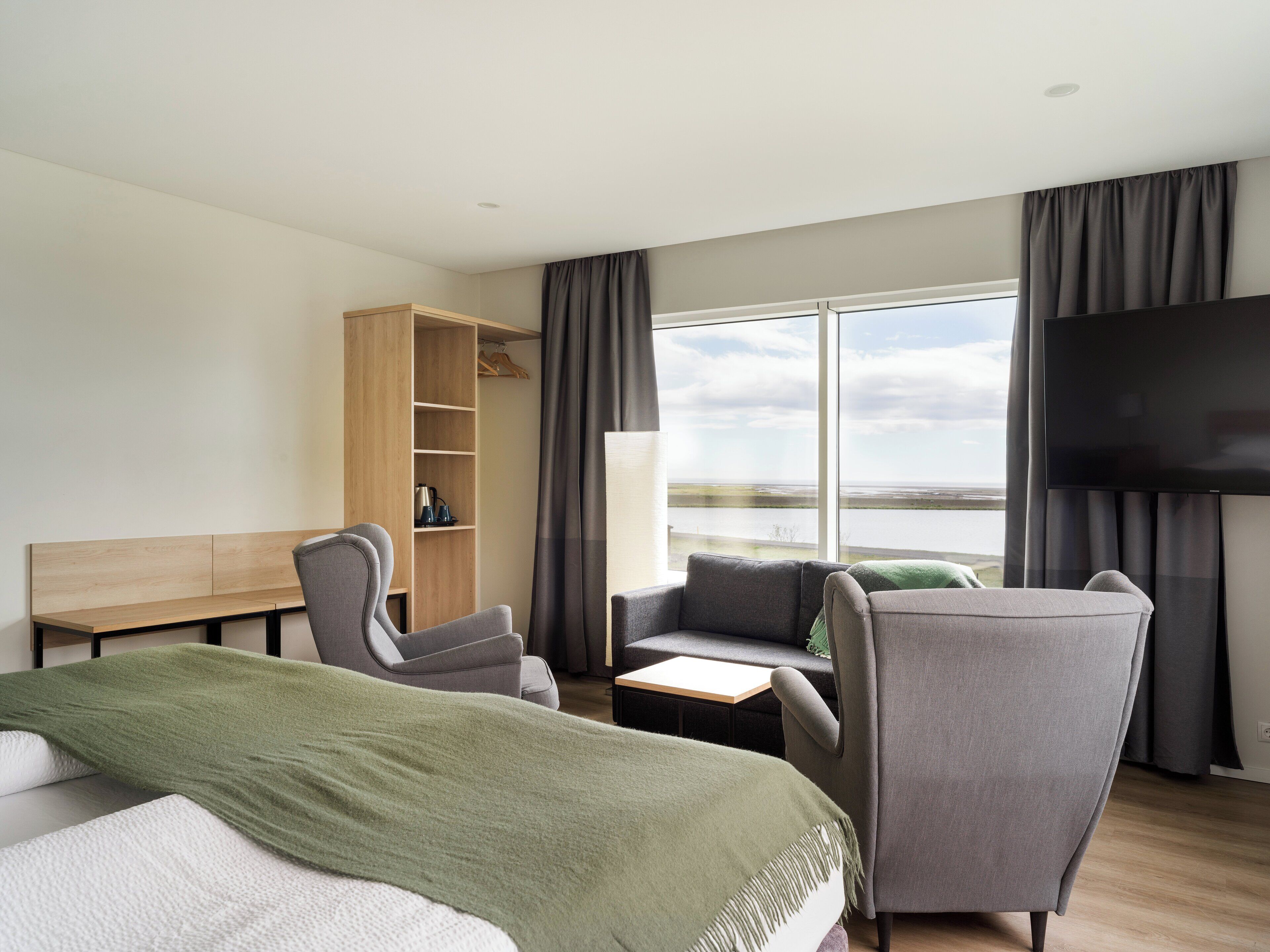 Quarto de hotel moderno com uma cama grande, duas poltronas, uma mesinha, um guarda-roupa de madeira, uma televisão e uma janela ampla com vista para o mar.