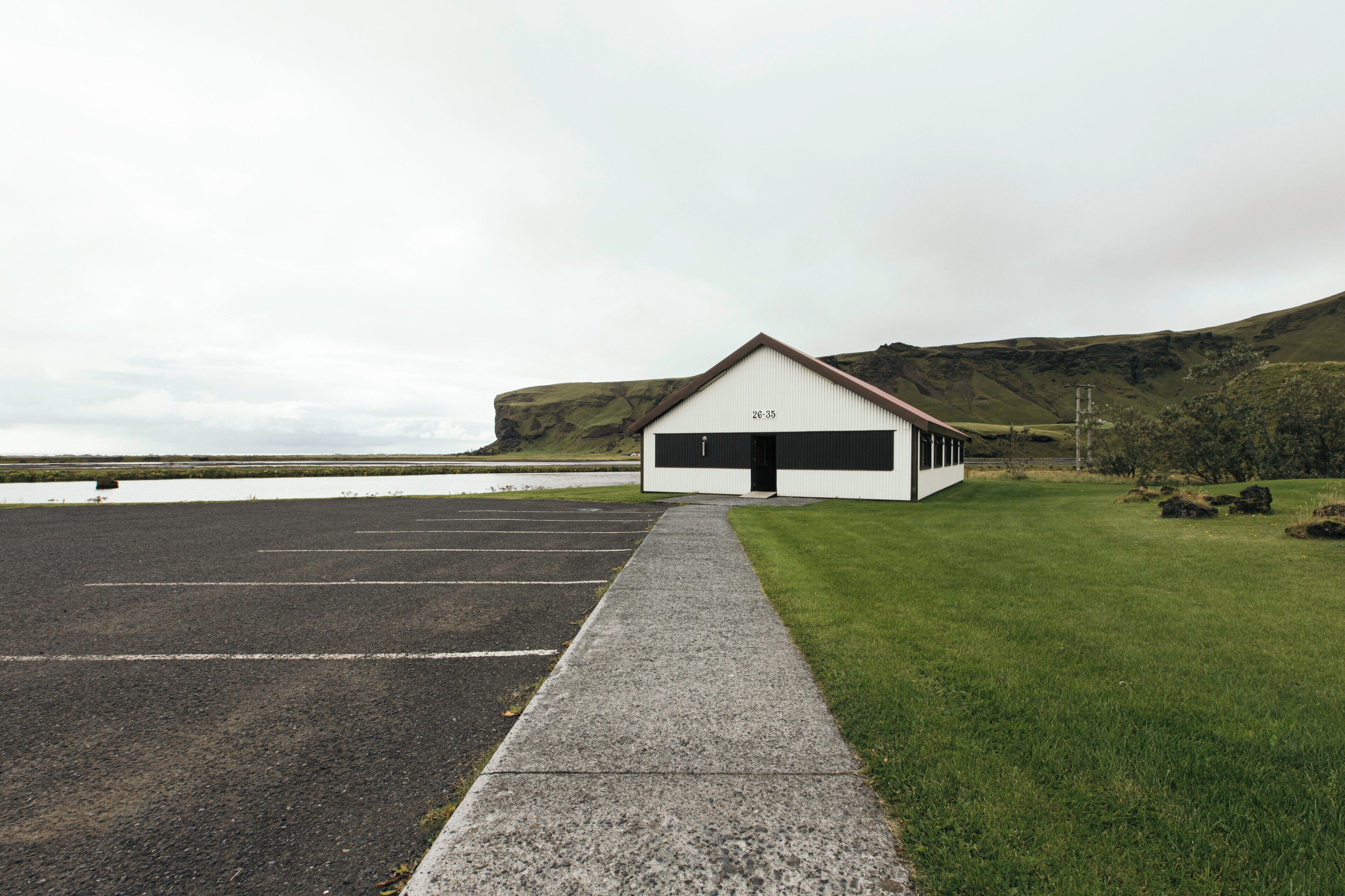 A white building with a black door and the number 26-35 on its facade, located next to a parking lot and a grassy area with a view of a body of water and hills in the background.