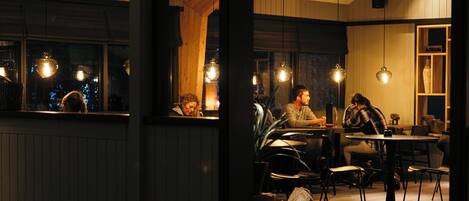 A cozy restaurant with wooden walls and hanging pendant lights. There are tables and chairs for dining.