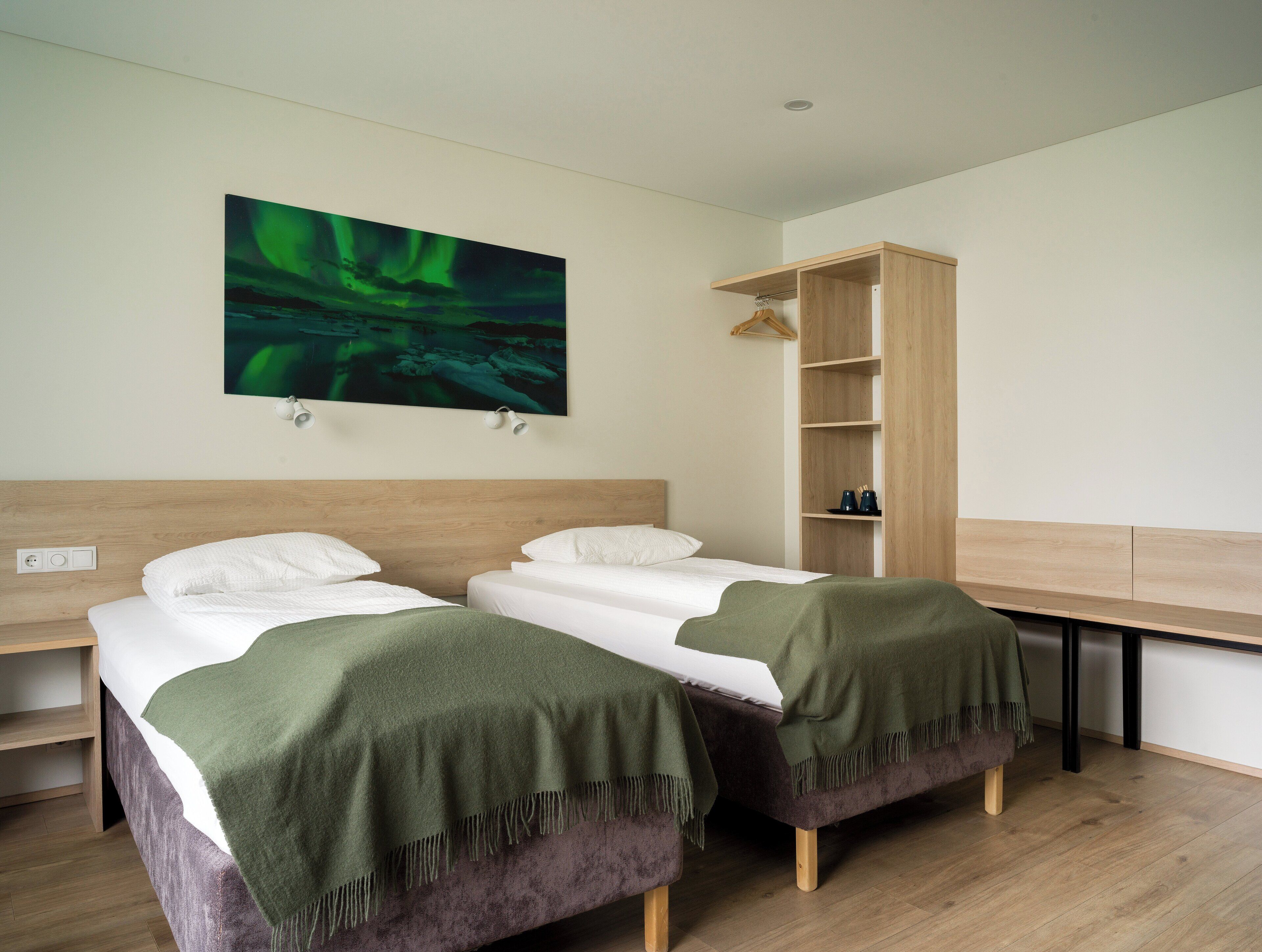 Hotelkamer met twee bedden, een houten hoofdeinde, een groene deken, een houten bureau en een schilderij van het noorderlicht.
