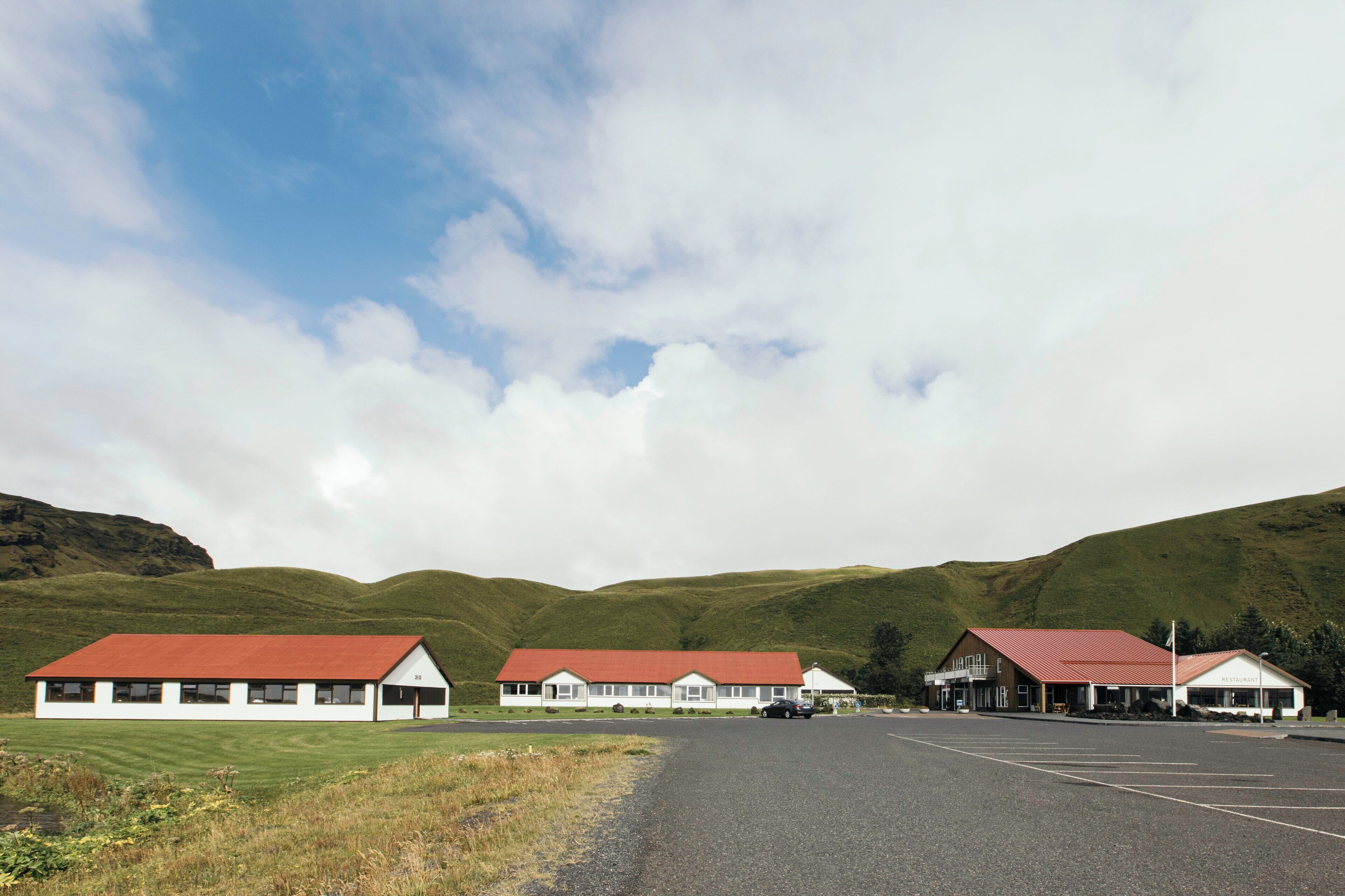 A row of white buildings with red roofs, a parking lot, and green hills in the background.