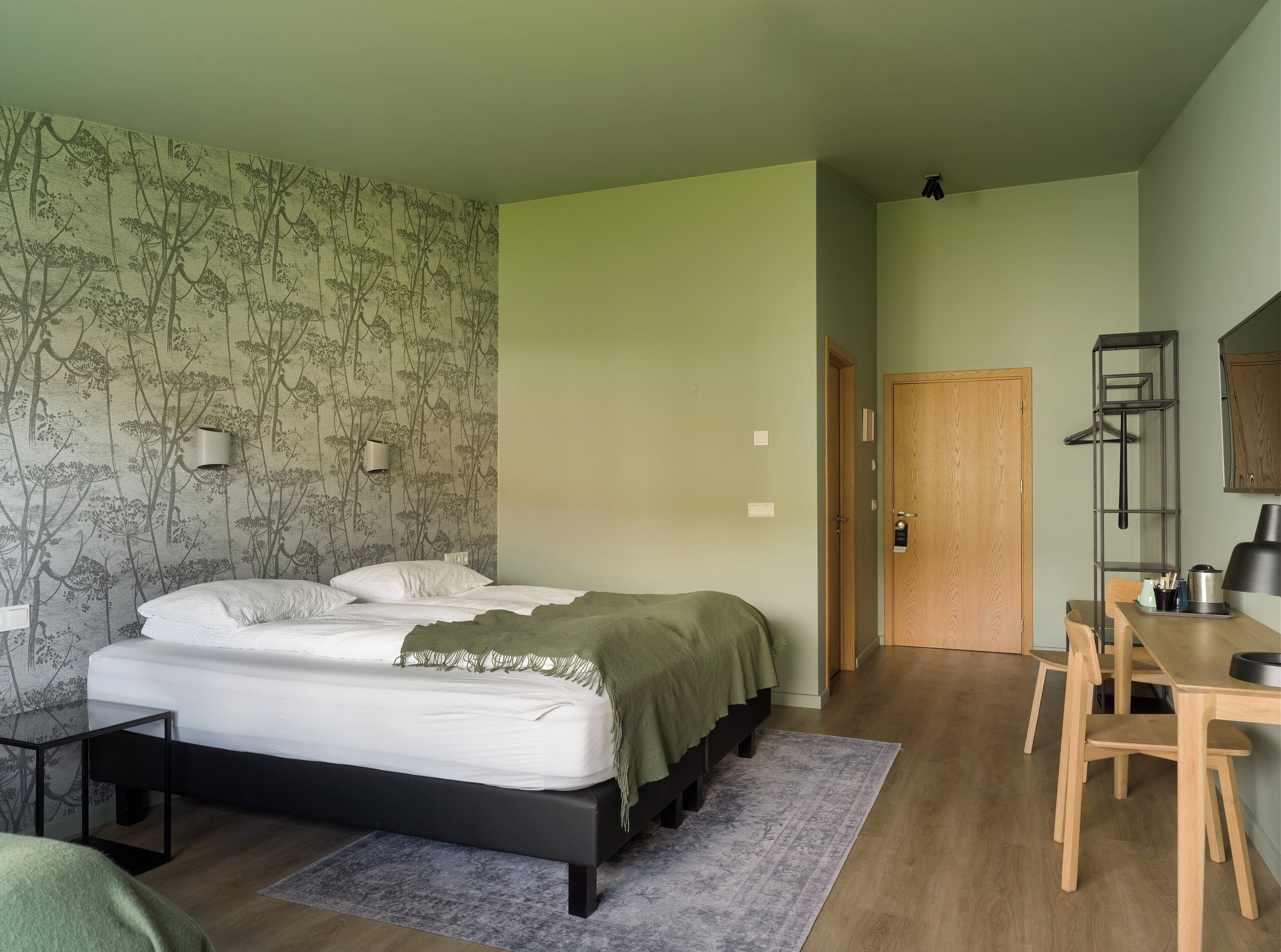 A bedroom with a bed, bedside table, and a desk with a chair. There is a door leading to another room.