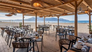 Un espace de restauration en plein air avec des tables et des chaises en bois, un toit de chaume et une vue sur la plage et les montagnes.