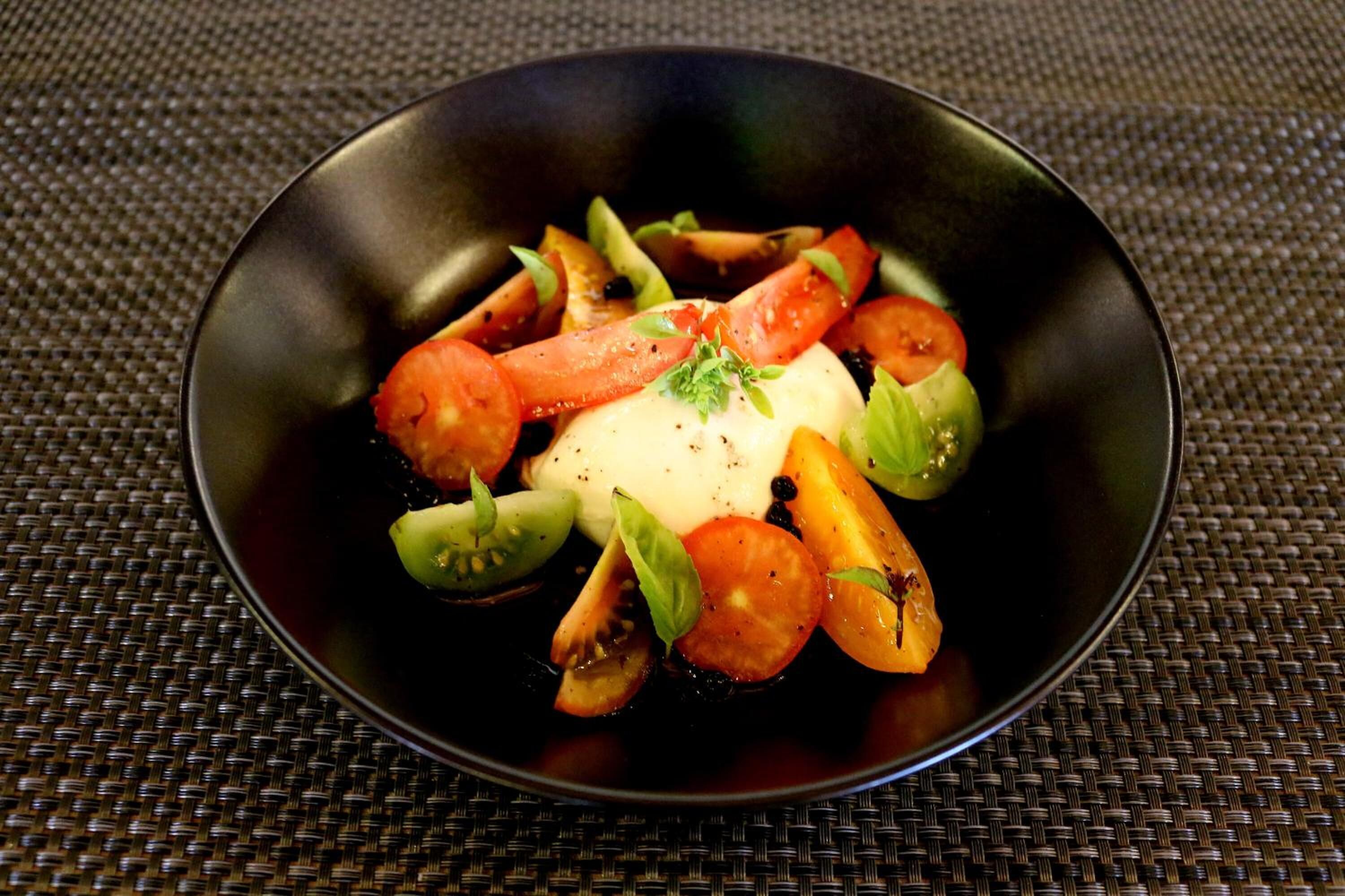 A black bowl containing a dish with tomatoes, basil, and a white sauce, placed on a woven mat.