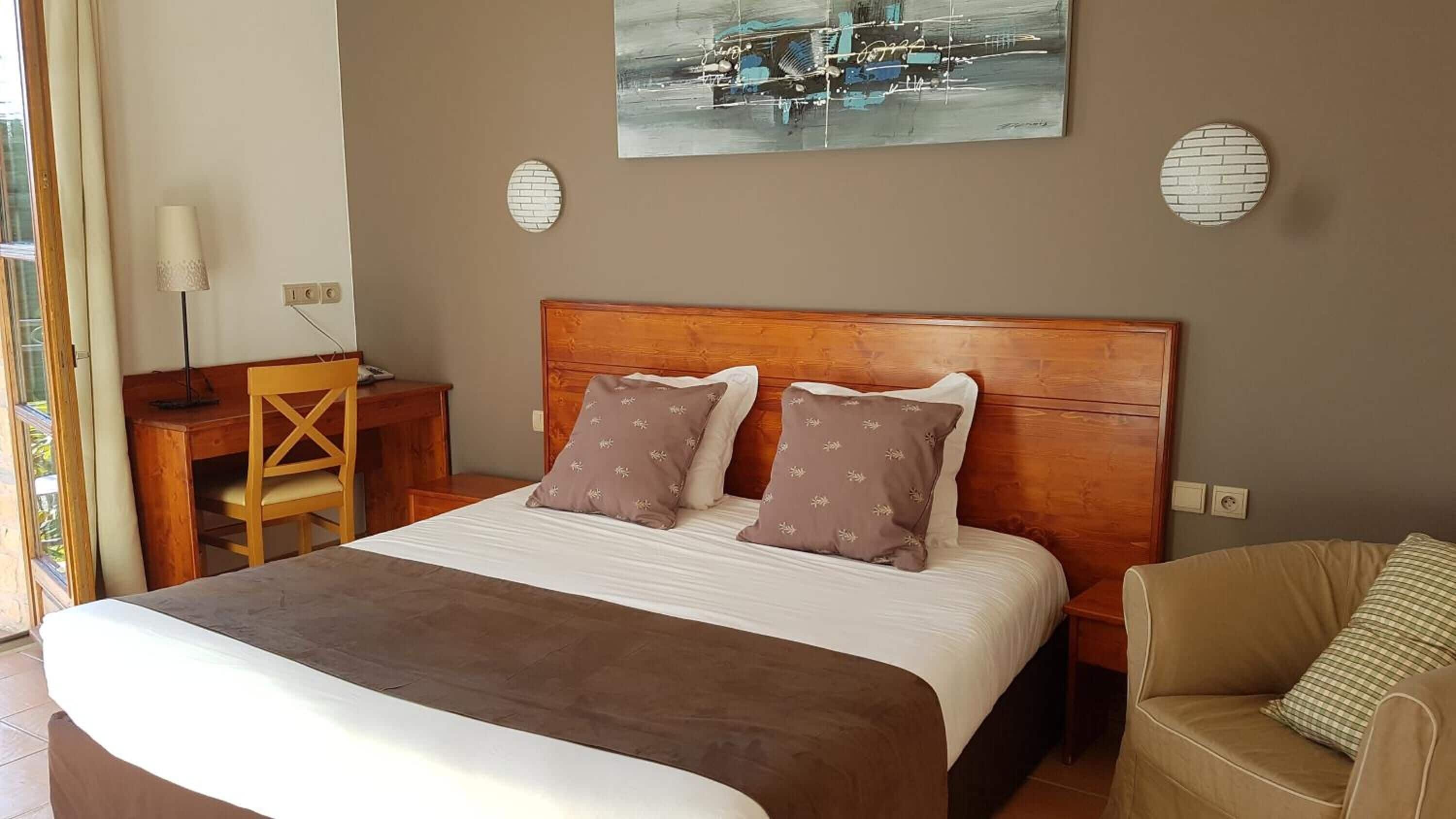 Habitación de hotel con cama de madera, dos almohadas, escritorio, silla, lámpara y un cuadro en la pared.