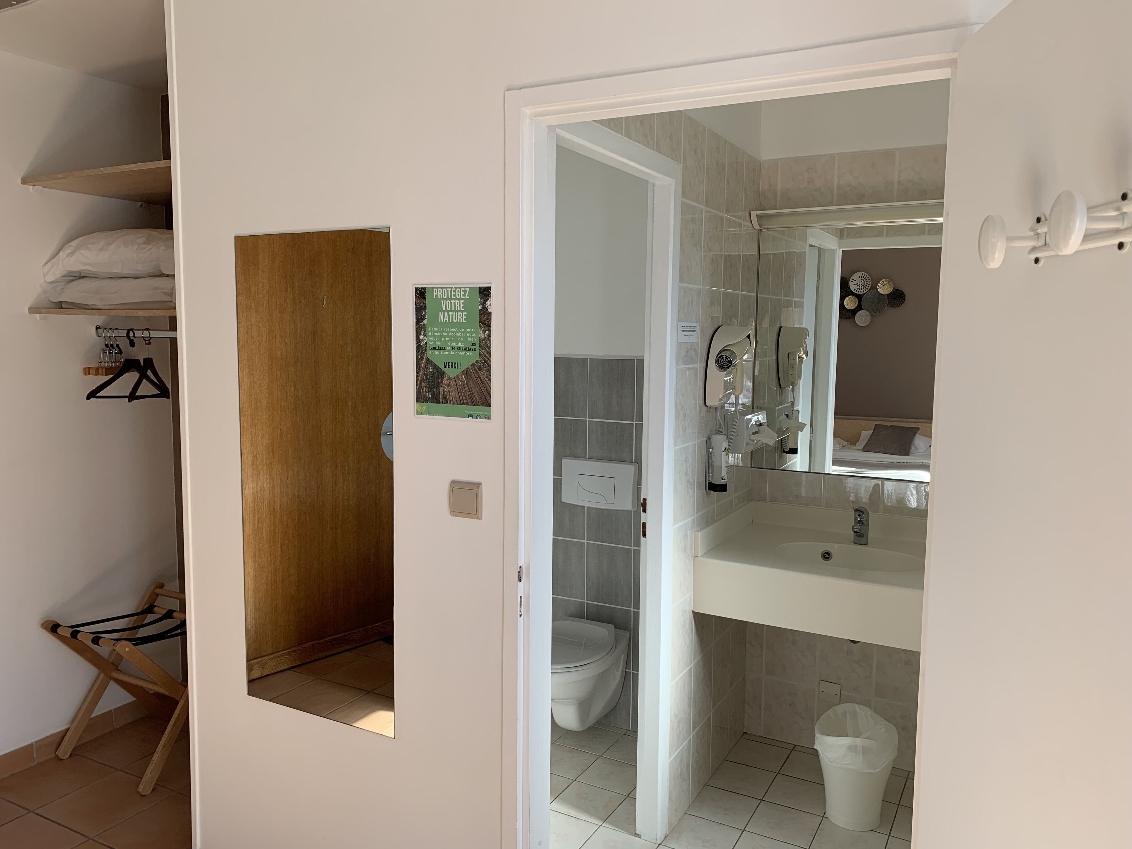 Baño con inodoro, lavamanos y espejo. Cuenta con zona de ducha con puerta de cristal y un perchero. Una puerta de madera da acceso a un armario con espacio para colgar ropa y un tendedero.