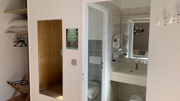 Baño con inodoro, lavamanos y espejo. Cuenta con zona de ducha con puerta de cristal y un perchero. Una puerta de madera da acceso a un armario con espacio para colgar ropa y un tendedero.