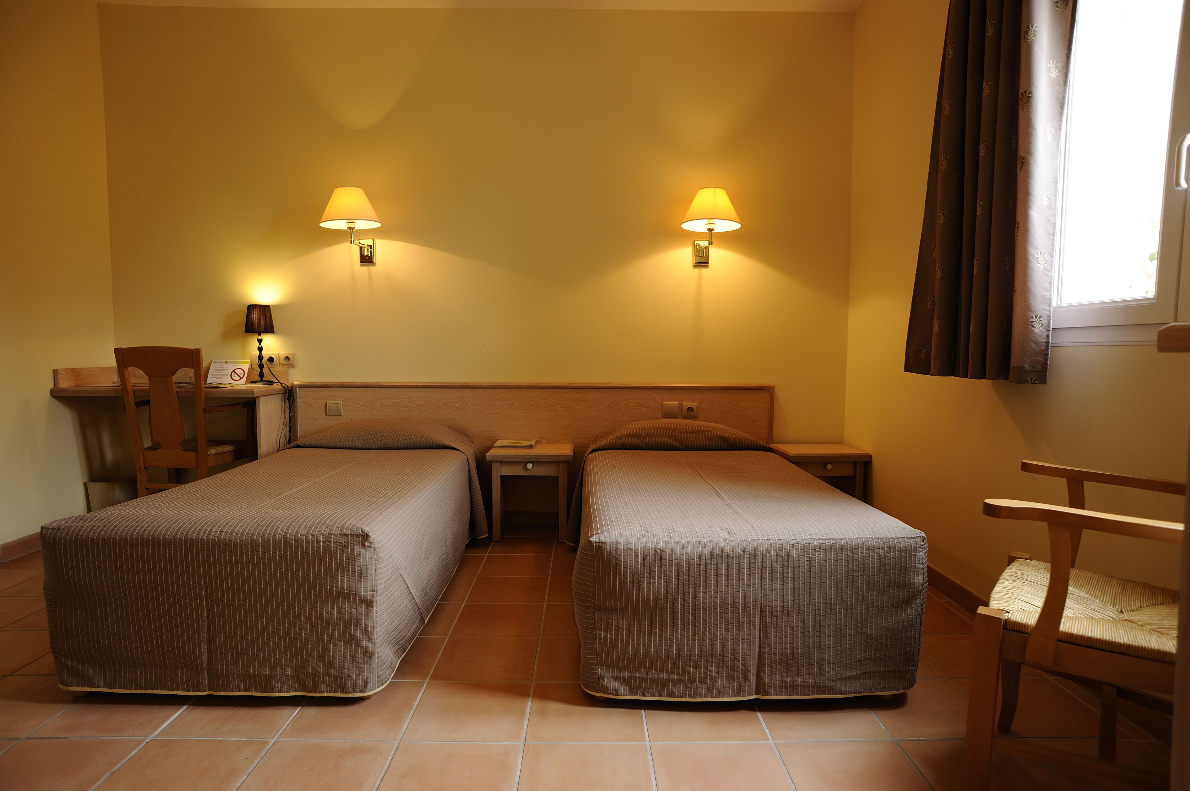 Une chambre d’hôtel avec deux lits simples, un bureau, une chaise et deux lampes fixées au mur.
