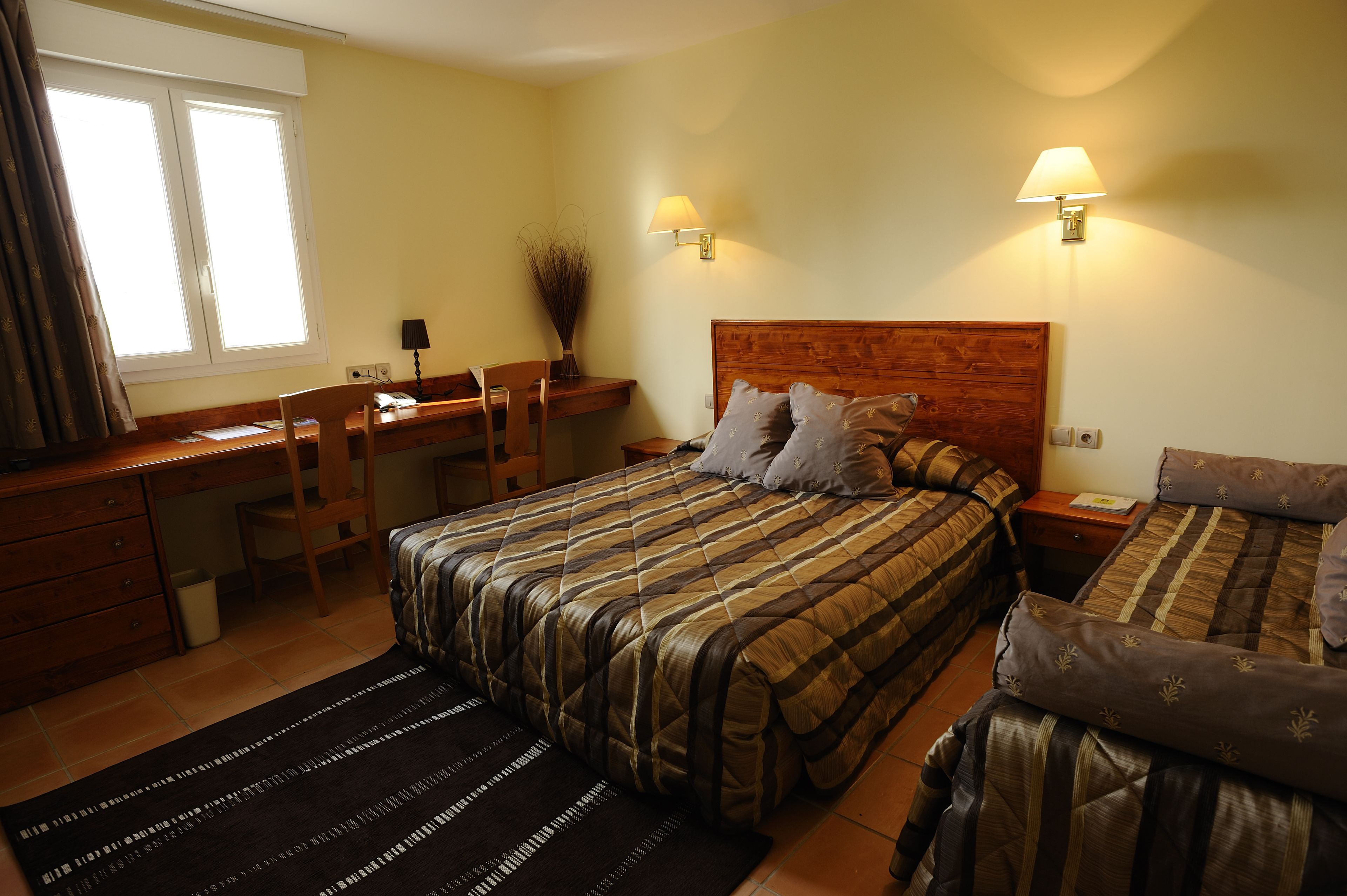A bedroom with a bed, a desk, and a chair. There are two bedside lamps and a window with curtains.
