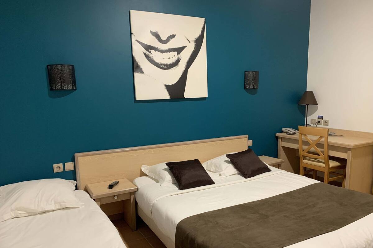 A hotel room with two beds, a desk, and a chair. There is a painting on the wall and a lamp on the desk.