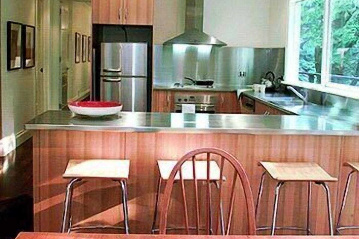 A modern kitchen with stainless steel appliances, a central island with bar stools, and a view of greenery through a window.