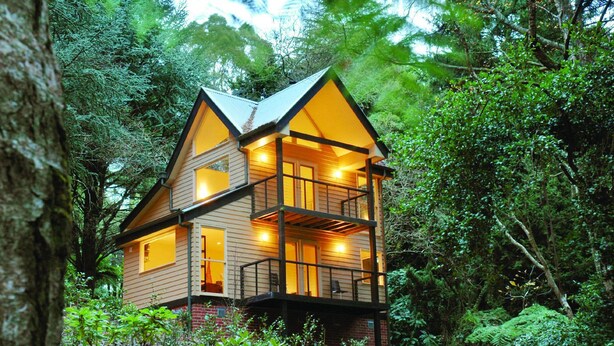 A two-story wooden cabin with a balcony, surrounded by dense forest.