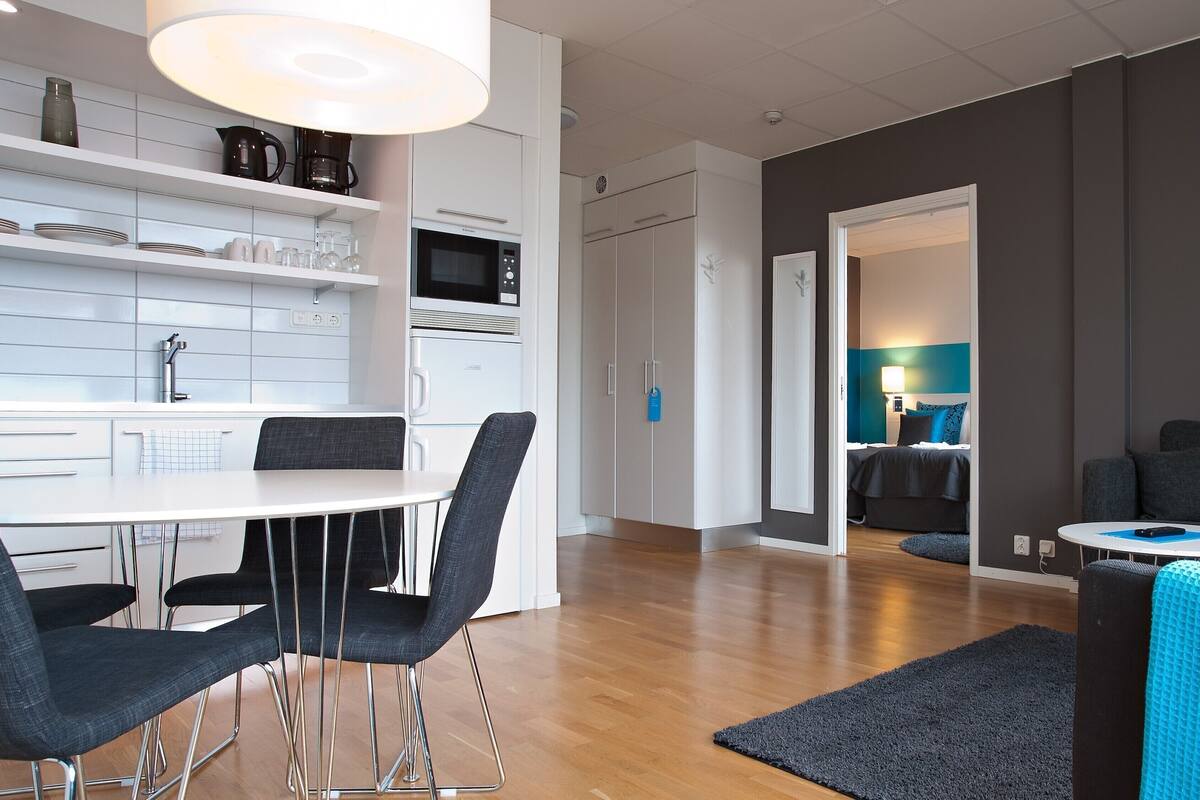 A modern kitchen with white cabinetry, a microwave, and a coffee maker. There is a dining area with a white table and black chairs. The room has wooden flooring and a gray rug.