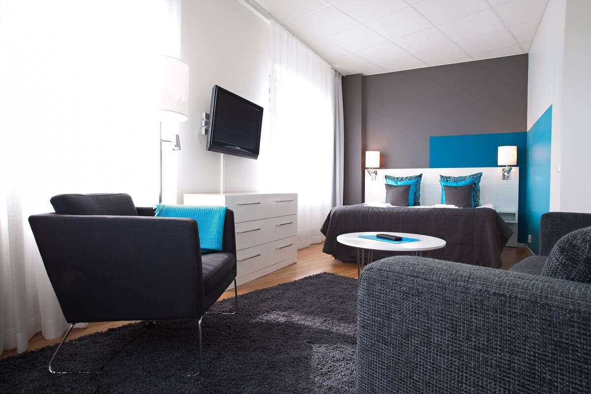 A modern hotel room with a flat-screen TV, a sofa, a bed with blue pillows, a bedside table, and a large window with curtains.