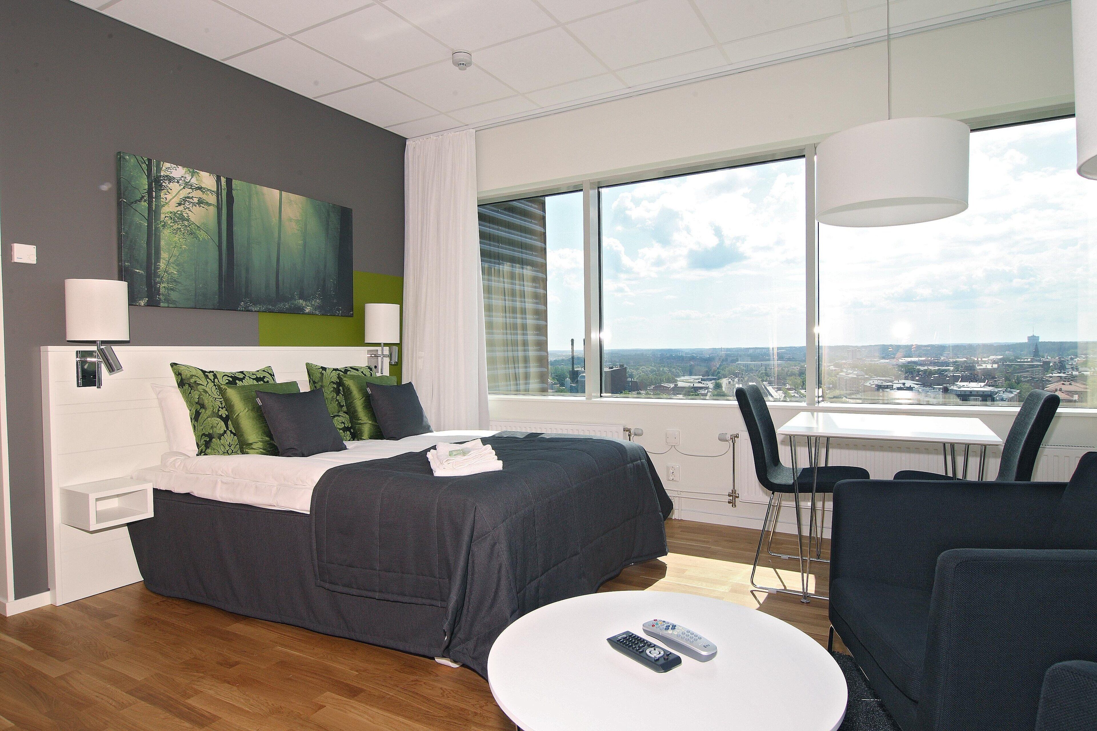 A modern hotel room with a large bed, a small table, and a view of the city through large windows.
