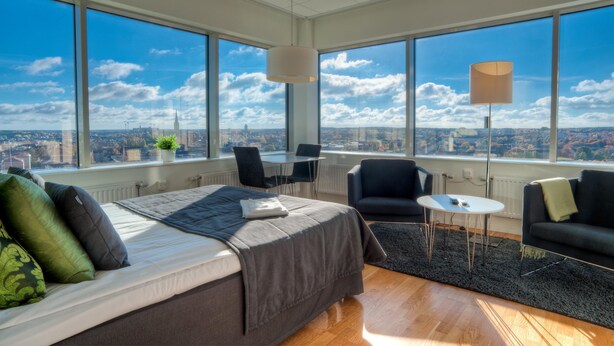 A modern hotel room with a large bed, two black armchairs, a small table, and a floor lamp. The room features large windows offering a city view. - Sky Hotel Apartments Tornet (Linkoping)