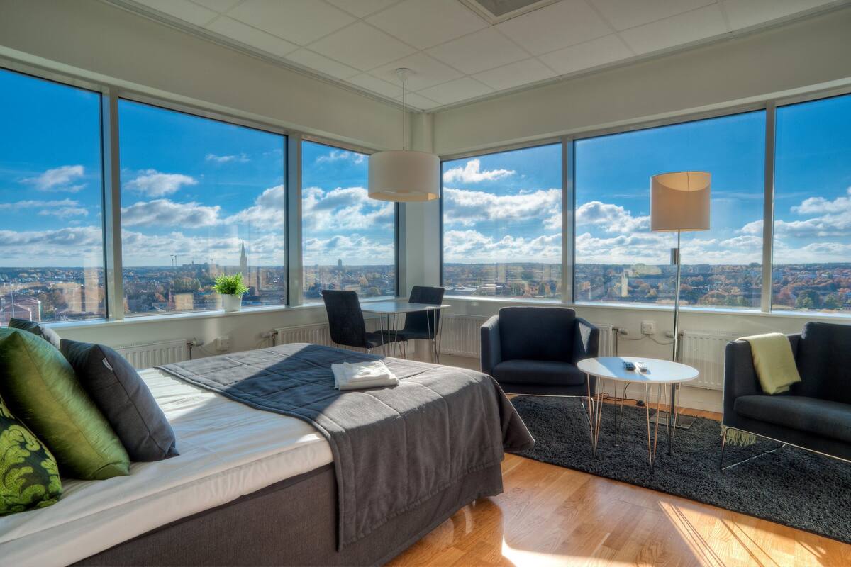 A modern hotel room with a large bed, two black armchairs, a small table, and a floor lamp. The room features large windows offering a city view.