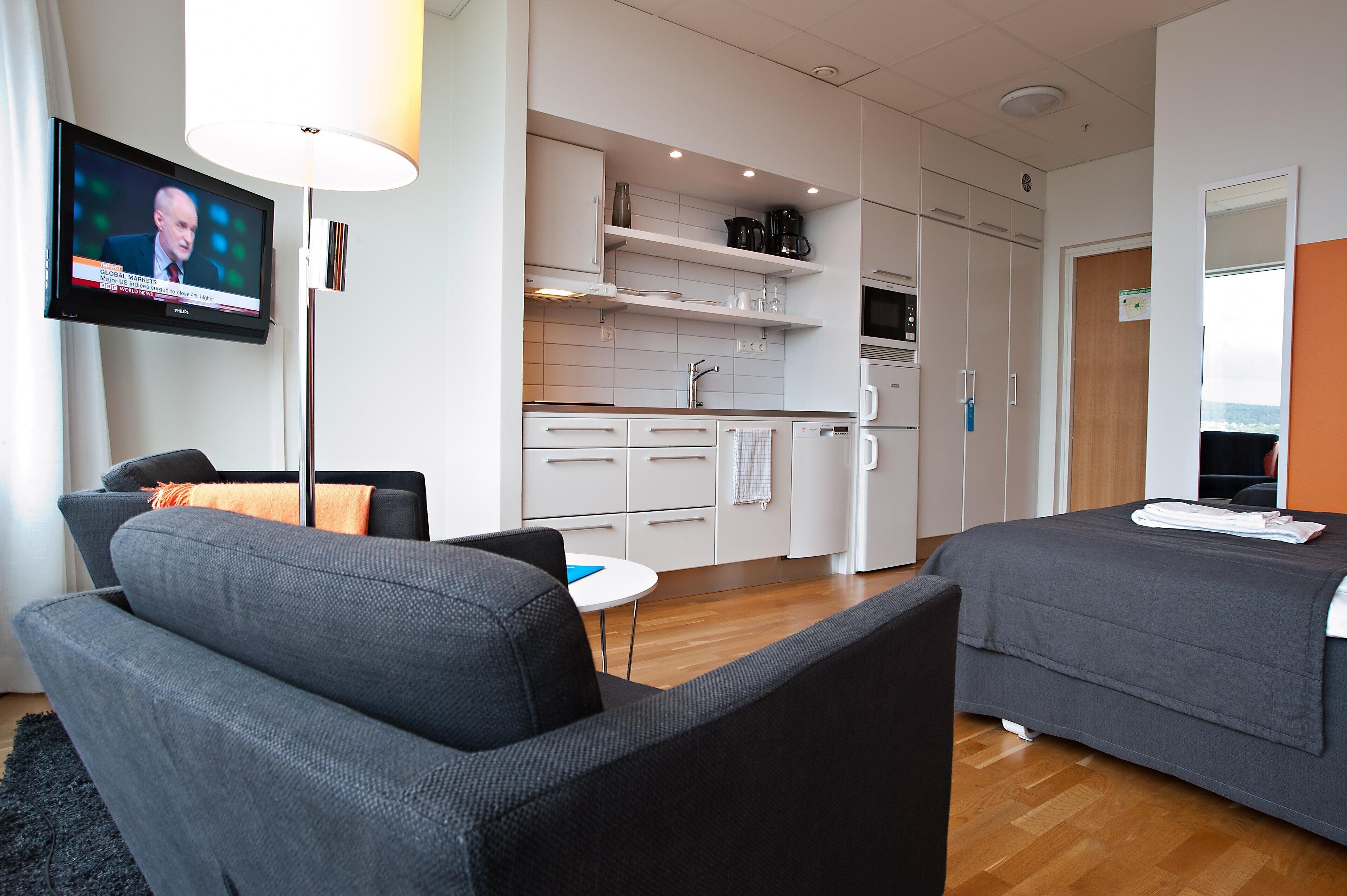Een moderne hotelkamer met een bank, een bed, een kitchenette en een televisie waarop een nieuwsuitzending draait.