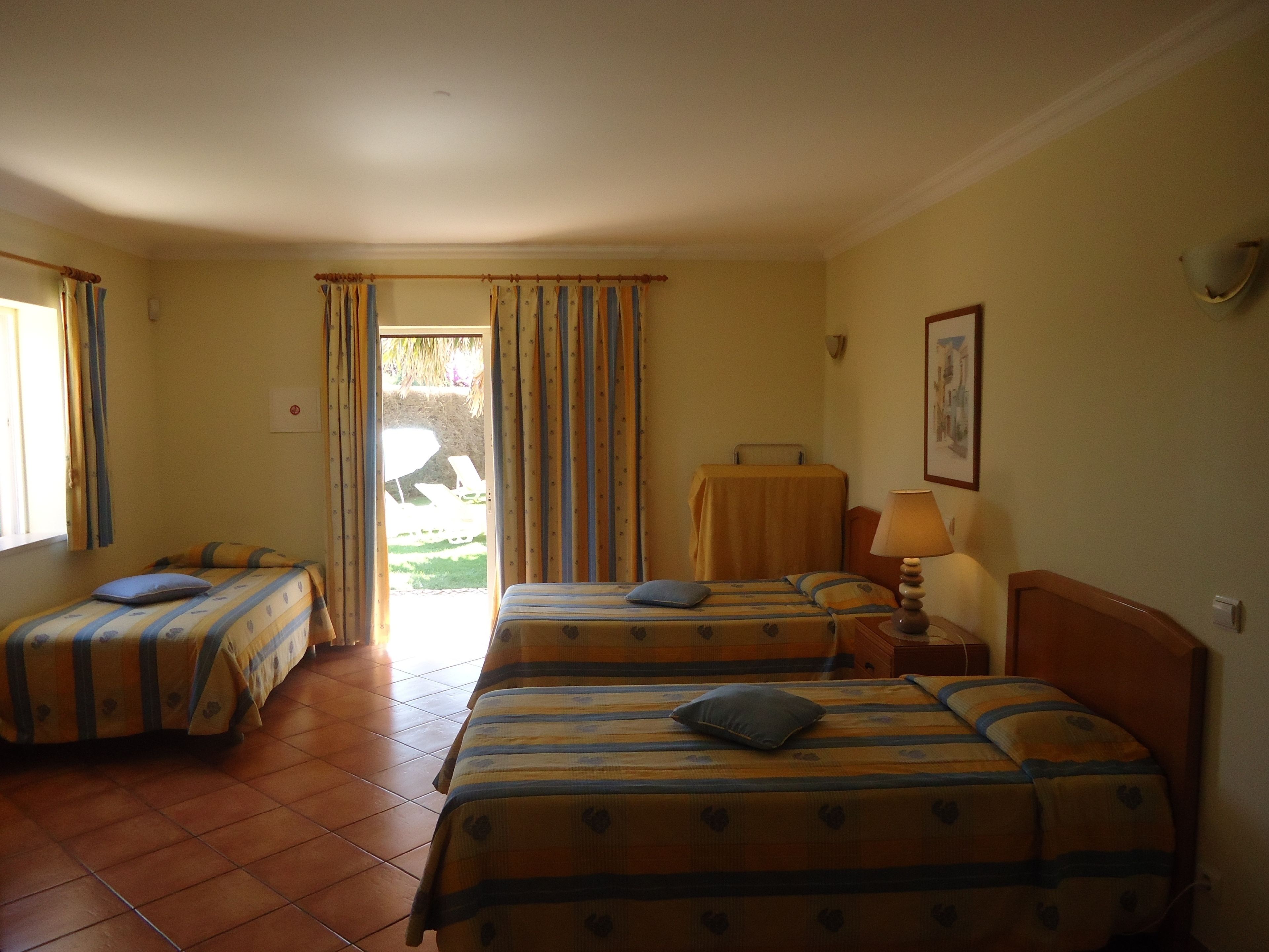 Una habitación de hotel con dos camas, una ventana, una puerta que da al exterior y una mesa con una lámpara.