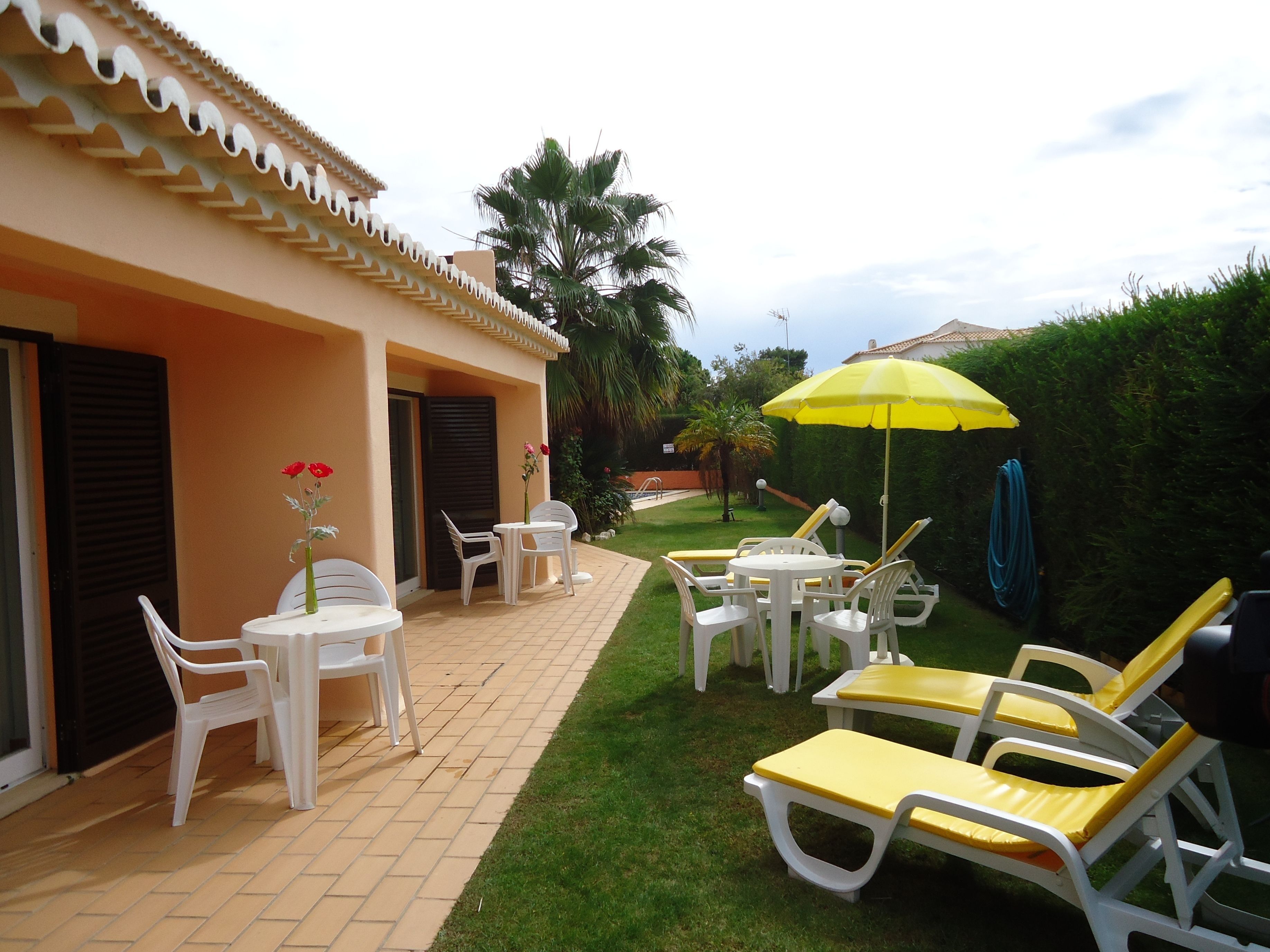 Un patio con sillas y mesas de plástico blanco, una sombrilla amarilla y una piscina al fondo.