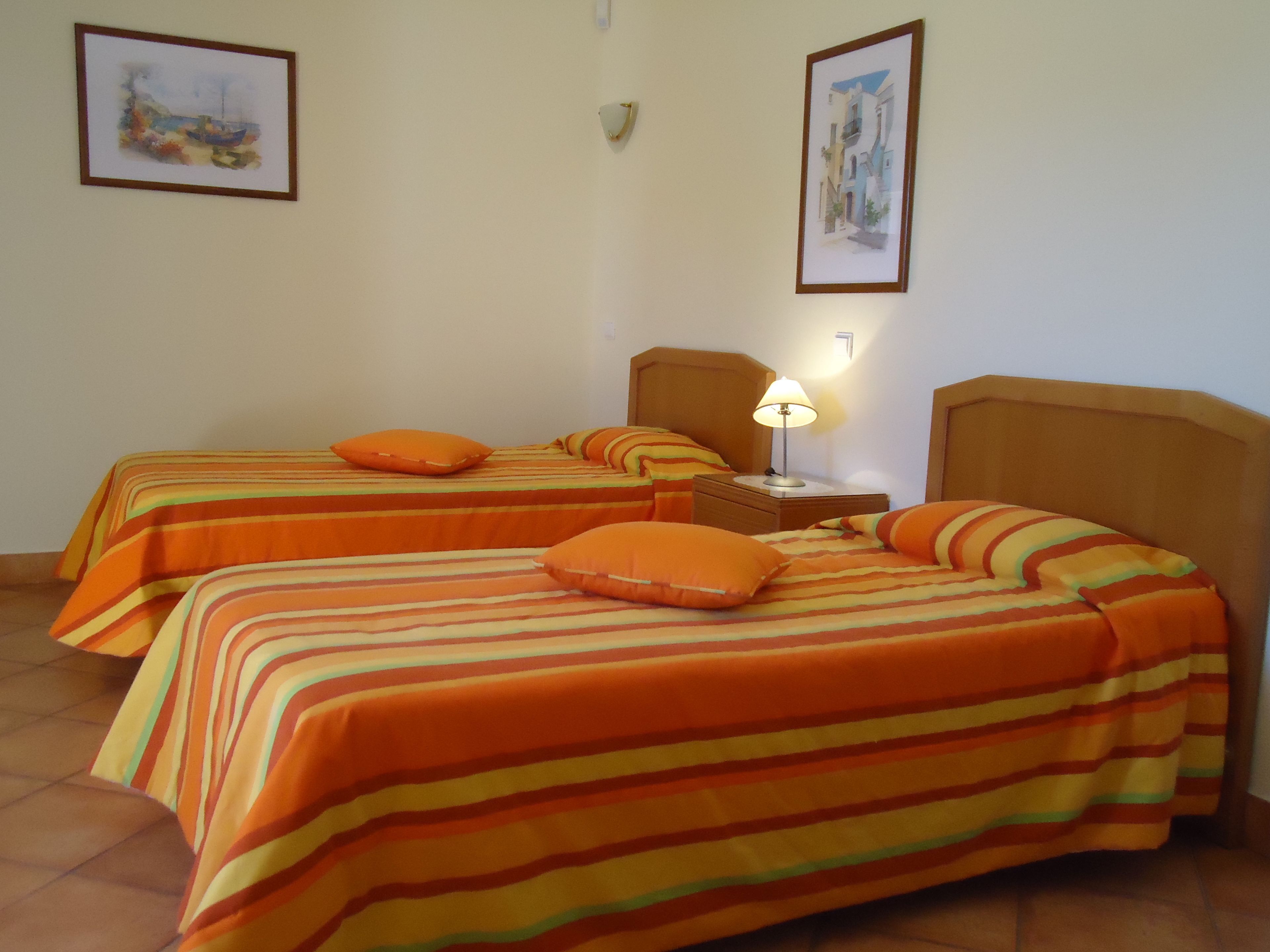 A hotel room with two beds, each with striped bedding and orange pillows. There is a nightstand with a lamp between the beds.