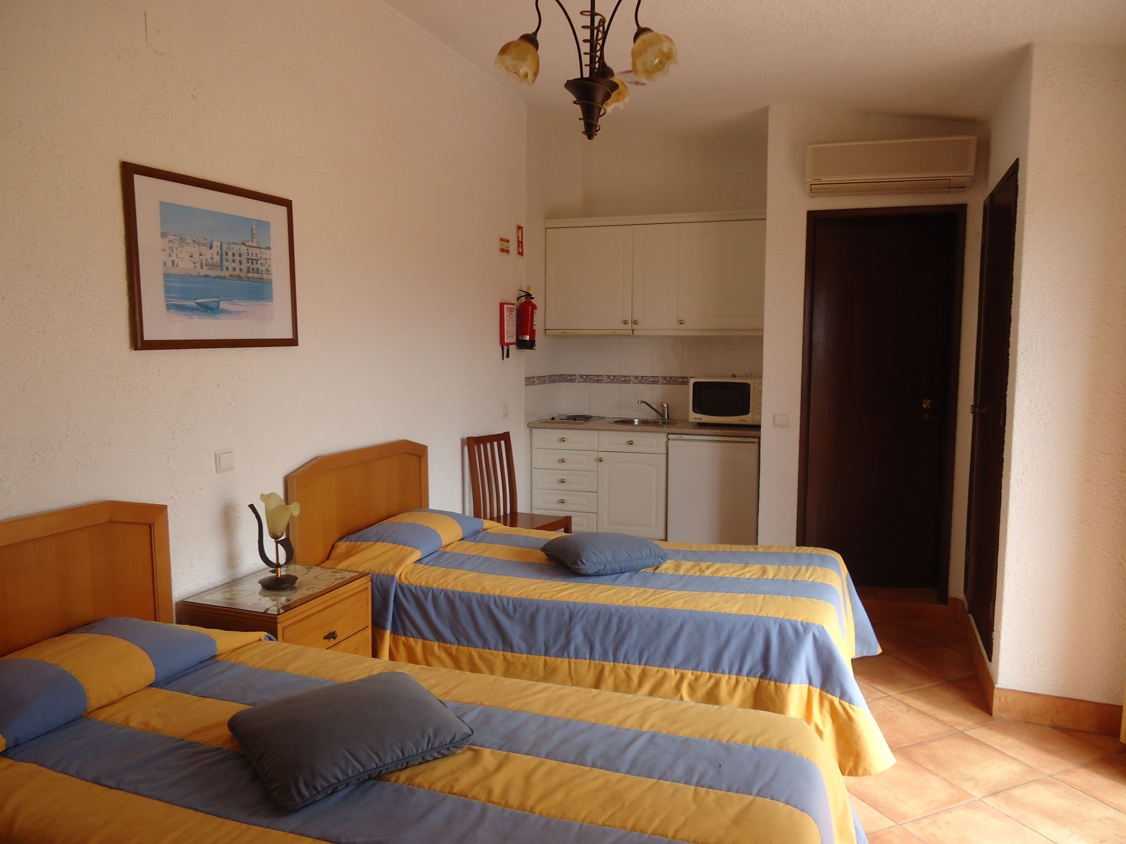 A bedroom with two beds, a small bedside table, a nightstand, a framed picture on the wall, a kitchen area with a microwave, and a door leading to another room.