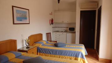 A bedroom with two beds, a small bedside table, a nightstand, a framed picture on the wall, a kitchen area with a microwave, and a door leading to another room.