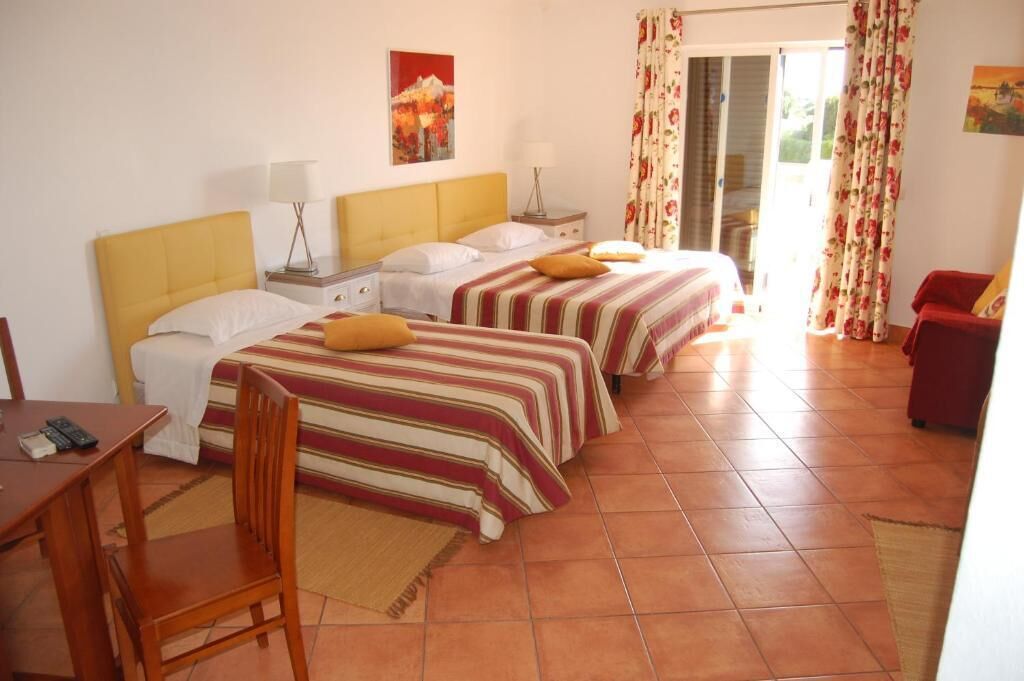 A hotel room with two beds, a desk, and a chair. There is a window with curtains, a painting on the wall, and a red chair in the corner.